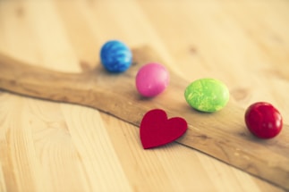 A wooden surface with five colorful Easter eggs scattered on it, including blue, pink, green, and red eggs. There is also a small red heart cutout positioned in front of the eggs.