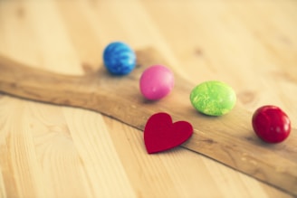 A wooden surface with five colorful Easter eggs scattered on it, including blue, pink, green, and red eggs. There is also a small red heart cutout positioned in front of the eggs.
