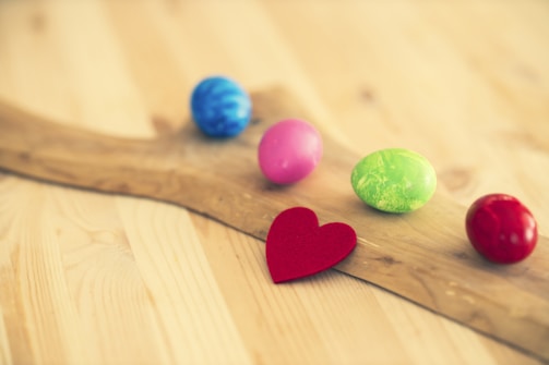 A wooden surface with five colorful Easter eggs scattered on it, including blue, pink, green, and red eggs. There is also a small red heart cutout positioned in front of the eggs.