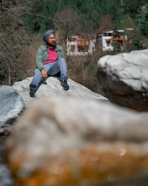 A man wearing a turban and casual clothing sits on a large rock in a natural setting. Behind him, there are trees and a house partially visible amongst the greenery, suggesting a serene outdoor environment.