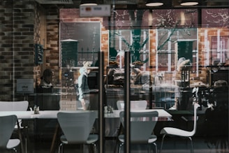 A cozy cafe with modern interior design featuring white chairs and tables against a backdrop of exposed brick walls. Chalkboard menus displaying items and prices are hanging on the walls. Reflections of trees and street life are visible through the large glass windows. People inside are engaged in various activities, such as ordering or preparing food.
