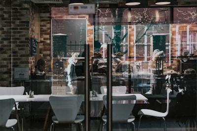 A cozy cafe with modern interior design featuring white chairs and tables against a backdrop of exposed brick walls. Chalkboard menus displaying items and prices are hanging on the walls. Reflections of trees and street life are visible through the large glass windows. People inside are engaged in various activities, such as ordering or preparing food.