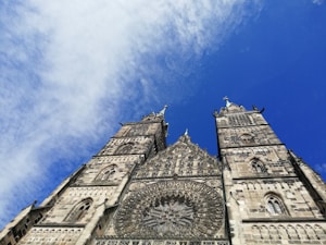 A grand Gothic-style cathedral with intricate stonework and towering spires set against a bright blue sky. The facade features elaborate carvings and a large circular stained glass window.