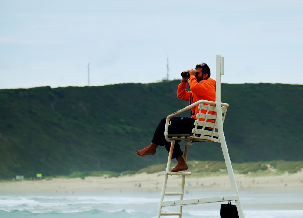 homme en chemise orange assis sur une chaise pliante en bois blanc sur la plage pendant la journée