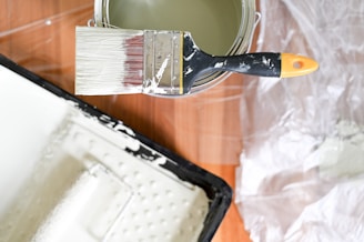 A paintbrush with white paint is resting on a paint can. There is a paint tray nearby on a wooden surface, and a plastic sheet is partially visible in the background.