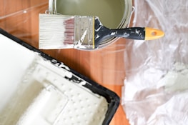 A paintbrush with white paint is resting on a paint can. There is a paint tray nearby on a wooden surface, and a plastic sheet is partially visible in the background.