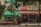 A small outdoor market shop covered with a Coca-Cola branded canopy in an urban setting. The shop appears to sell a variety of goods and is partially obscured by hanging green plants. The environment seems aged and slightly worn, evoking a sense of nostalgia.