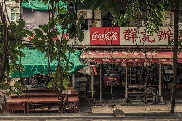 A small outdoor market shop covered with a Coca-Cola branded canopy in an urban setting. The shop appears to sell a variety of goods and is partially obscured by hanging green plants. The environment seems aged and slightly worn, evoking a sense of nostalgia.