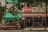A small outdoor market shop covered with a Coca-Cola branded canopy in an urban setting. The shop appears to sell a variety of goods and is partially obscured by hanging green plants. The environment seems aged and slightly worn, evoking a sense of nostalgia.