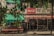A small outdoor market shop covered with a Coca-Cola branded canopy in an urban setting. The shop appears to sell a variety of goods and is partially obscured by hanging green plants. The environment seems aged and slightly worn, evoking a sense of nostalgia.