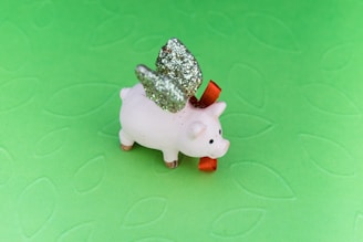 A small ceramic pig figure with glittery silver wings and a red bow around its neck is set against a textured light green background.