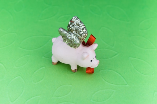 A small ceramic pig figure with glittery silver wings and a red bow around its neck is set against a textured light green background.
