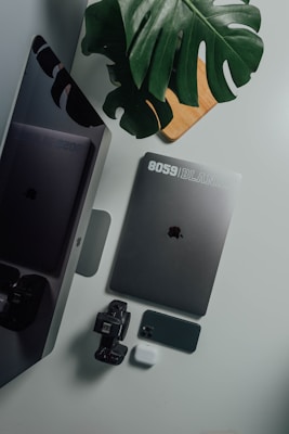 A collection of tech gadgets including a laptop, smartphone, smartwatch, and wireless earbuds placed on a white surface. A large green leaf partially covers the items, adding a touch of nature to the arrangement. A wooden board is partially visible underneath the leaf.