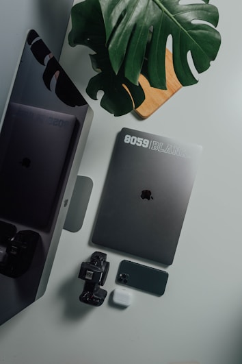 A collection of tech gadgets including a laptop, smartphone, smartwatch, and wireless earbuds placed on a white surface. A large green leaf partially covers the items, adding a touch of nature to the arrangement. A wooden board is partially visible underneath the leaf.