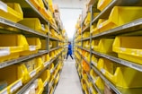 A person in a blue uniform stands in a warehouse aisle, surrounded by shelves filled with yellow plastic bins. The bins are organized and labeled, containing various items, while the industrial lighting provides a bright environment.