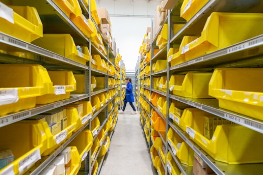 A person in a blue uniform stands in a warehouse aisle, surrounded by shelves filled with yellow plastic bins. The bins are organized and labeled, containing various items, while the industrial lighting provides a bright environment.