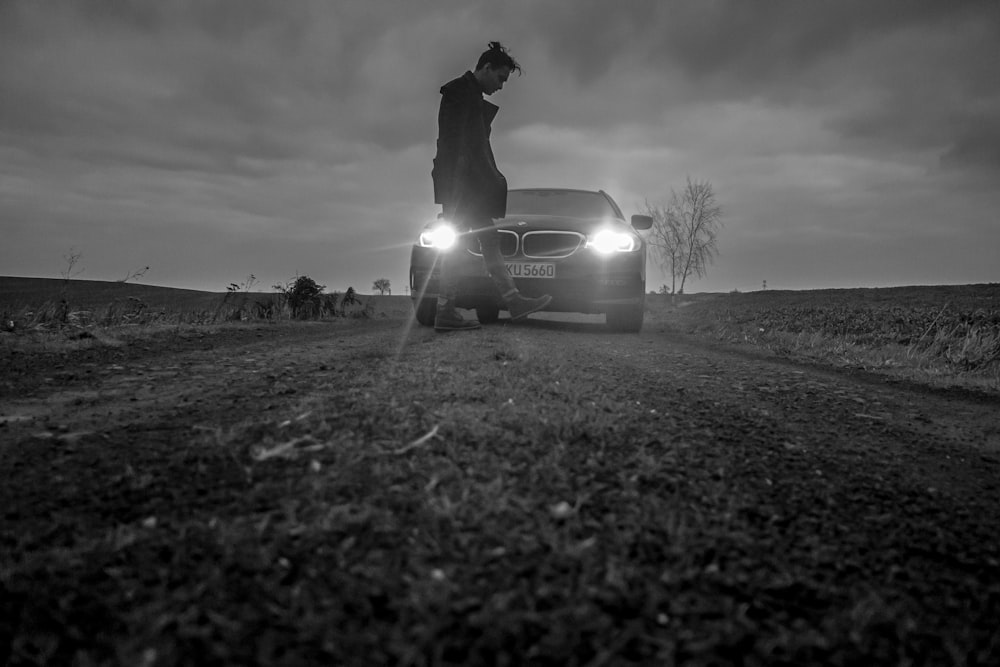 grayscale photo of man sitting on car hood