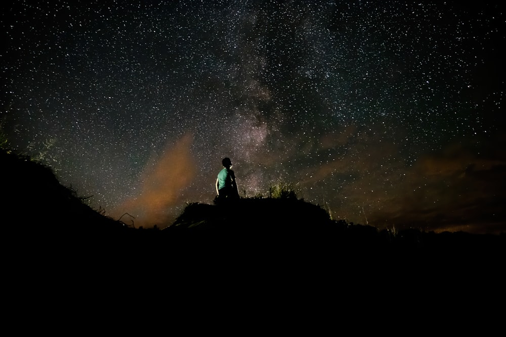 silhouette of man sitting on rock under starry night