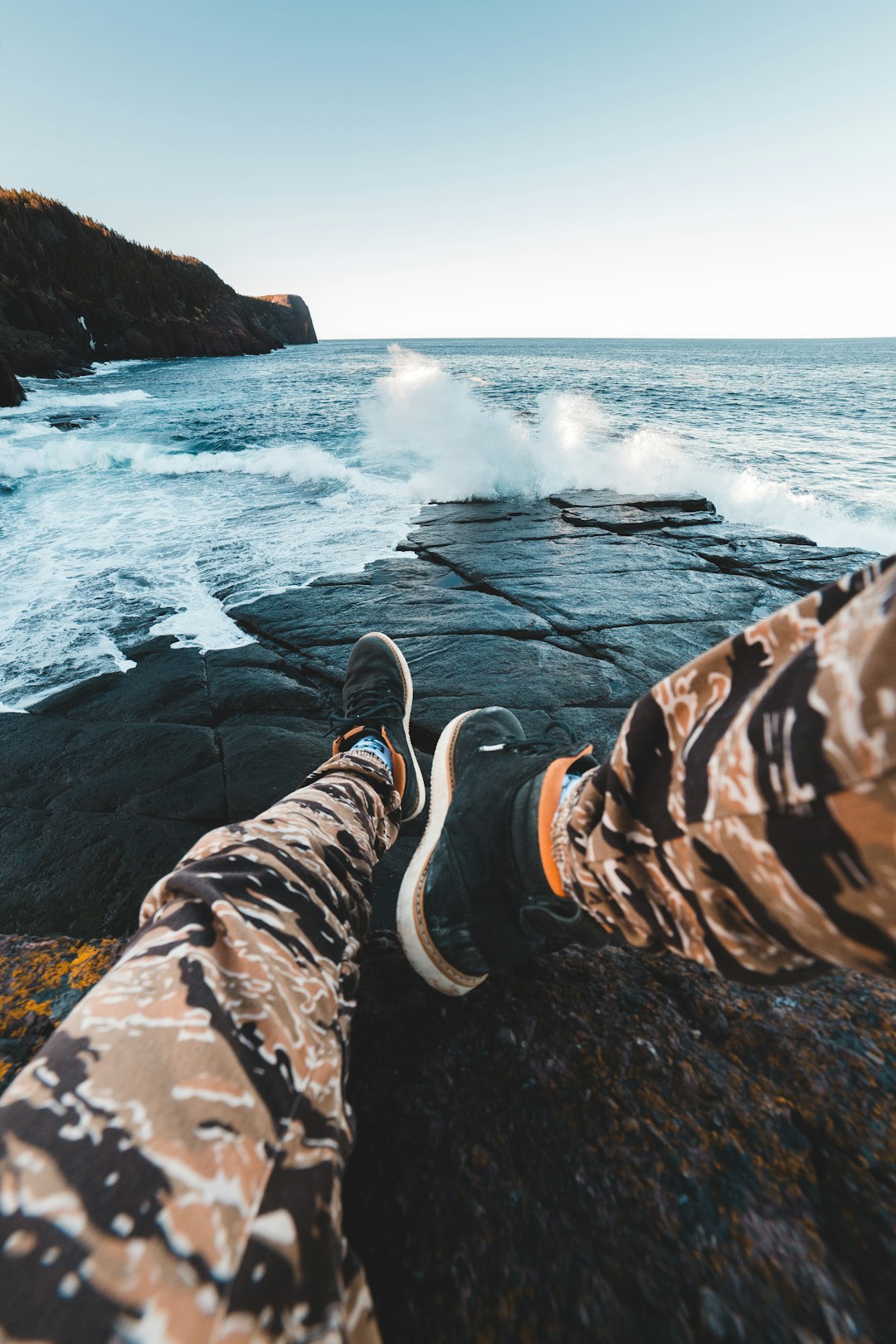 person in brown pants and black shoes sitting on rock formation near sea waves crashing on during