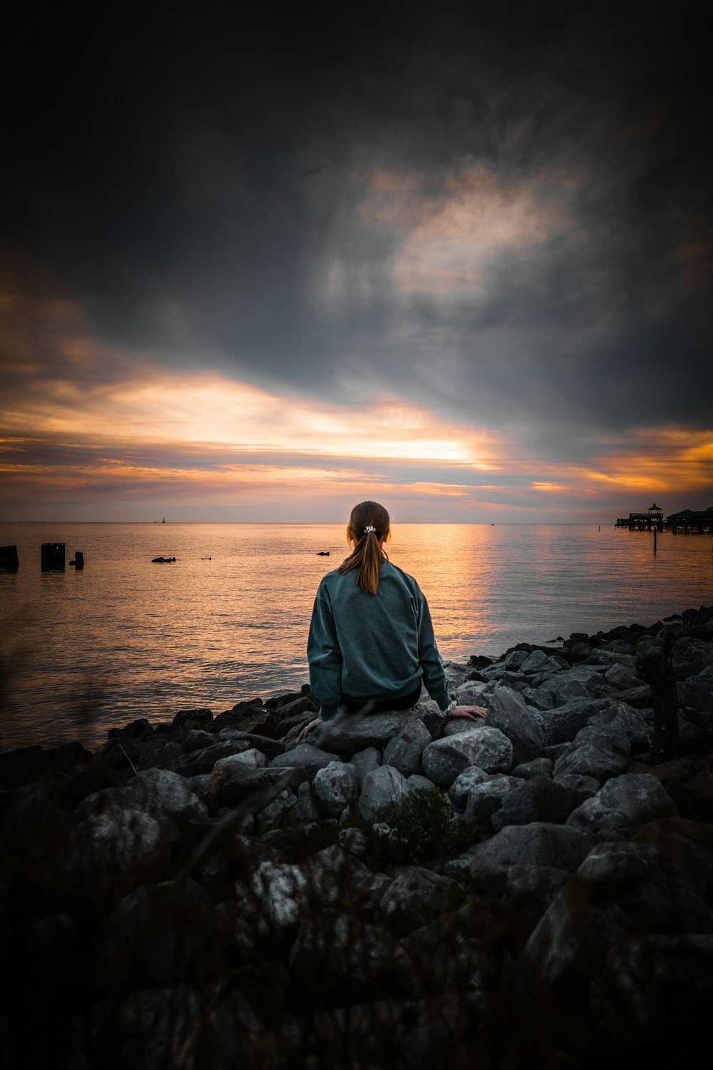 푸른 긴 소매 셔츠를 입은 여자는 일몰 동안 수역 근처의 회색 바위에 앉아 있다