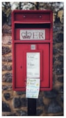 A traditional red postbox with a royal insignia and 'ER' marking is built into a stone wall. Below, there is a sign indicating collection times for mail. A handwritten note attached to the postbox thanks the postman for continuing to collect mail.