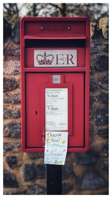 A traditional red postbox with a royal insignia and 'ER' marking is built into a stone wall. Below, there is a sign indicating collection times for mail. A handwritten note attached to the postbox thanks the postman for continuing to collect mail.
