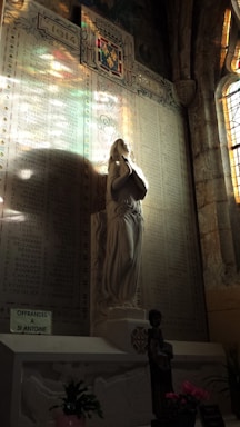 A detailed wall memorial with an engraved list of names, illuminated by colorful stained glass light. A stone statue of a person in a praying posture stands in front. Below the statue are various flowers and a small sign referring to offerings for a saint.