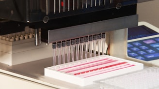 A laboratory instrument with multiple pipettes aligned over a tray is dispensing liquid into small wells. The background features part of a machine interface with buttons.