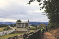 A rural landscape featuring an old stone granary with a wooden structure on top, along a winding road that leads into the distance. The background shows hills under a cloudy sky. Two people dressed for a hike are visible on a dirt path beside a stone wall, surrounded by trees and dry grass.
