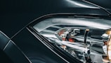Close-up view of a car headlight with intricate design, featuring reflective surfaces and sleek lines, with part of the vehicle body visible in metallic dark color. The word 'BMW' is embossed near the headlight.