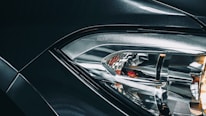 Close-up view of a car headlight with intricate design, featuring reflective surfaces and sleek lines, with part of the vehicle body visible in metallic dark color. The word 'BMW' is embossed near the headlight.