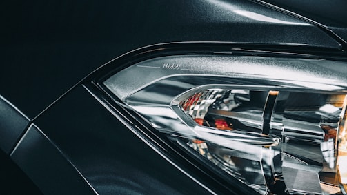 Close-up view of a car headlight with intricate design, featuring reflective surfaces and sleek lines, with part of the vehicle body visible in metallic dark color. The word 'BMW' is embossed near the headlight.