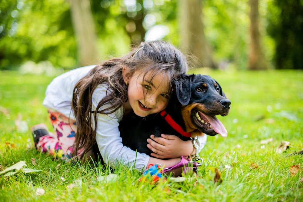 girl in pink jacket playing with black and brown short coated dog on green grass field