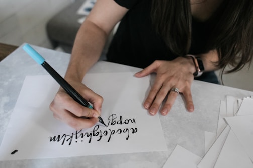 A person is practicing calligraphy on a white sheet of paper using a blue and black dual-tip marker. The writing appears artistic and smooth. The person is wearing a black shirt, and there is a watch on the wrist. Various blank sheets are scattered nearby on the table.