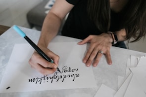 A person is practicing calligraphy on a white sheet of paper using a blue and black dual-tip marker. The writing appears artistic and smooth. The person is wearing a black shirt, and there is a watch on the wrist. Various blank sheets are scattered nearby on the table.