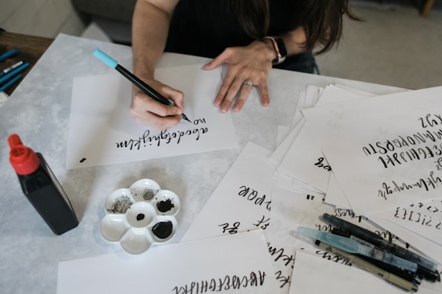 A person is practicing calligraphy with a black ink pen on a sheet of paper. Beside them, there are several other sheets with calligraphy writing and tools such as pens, brushes, a palette with black ink, and a bottle of ink.