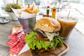 A wooden tray holds a large burger with a shiny, sesame seed bun. The burger is filled with lettuce, tomato, grilled onions, and melted cheese. Nearby, a metal cup contains French fries, and there are several small packets of tomato ketchup. In the background, there are condiment bottles and a glass of iced coffee.