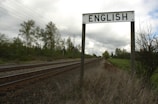 A railway track extends into the distance bordered by grassy areas and trees under a cloudy sky. A signpost prominently displays the word 'ENGLISH' above the tracks.