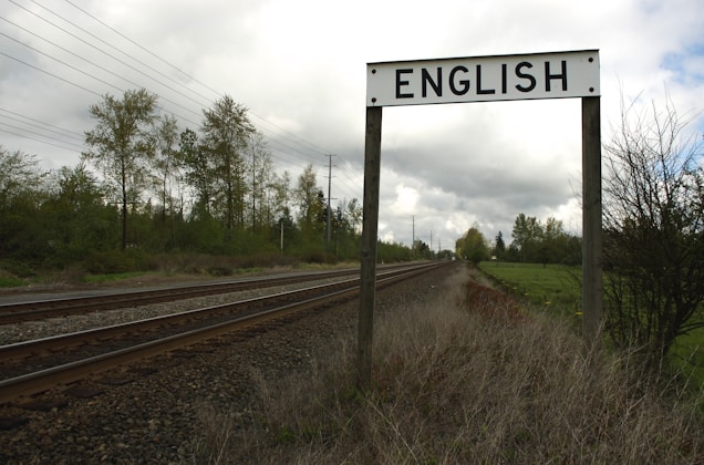 A railway track extends into the distance bordered by grassy areas and trees under a cloudy sky. A signpost prominently displays the word 'ENGLISH' above the tracks.