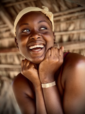 A person with a joyful expression, smiling widely and holding their hands near their face while wearing a light-colored headscarf. The background consists of a textured, natural material, possibly straw or wood, creating a warm and rustic atmosphere.