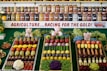 A colorful display of fruits and vegetables is organized in neat rows on three trays, surrounded by greenery and small stuffed animal figurines. Behind the display, shelves are filled with jars containing preserves and pickled goods. A banner with the text 'AGRICULTURE...RACING FOR THE GOLD!' is prominently featured.