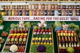 A colorful display of fruits and vegetables is organized in neat rows on three trays, surrounded by greenery and small stuffed animal figurines. Behind the display, shelves are filled with jars containing preserves and pickled goods. A banner with the text 'AGRICULTURE...RACING FOR THE GOLD!' is prominently featured.