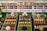 A colorful display of fruits and vegetables is organized in neat rows on three trays, surrounded by greenery and small stuffed animal figurines. Behind the display, shelves are filled with jars containing preserves and pickled goods. A banner with the text 'AGRICULTURE...RACING FOR THE GOLD!' is prominently featured.