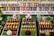 A colorful display of fruits and vegetables is organized in neat rows on three trays, surrounded by greenery and small stuffed animal figurines. Behind the display, shelves are filled with jars containing preserves and pickled goods. A banner with the text 'AGRICULTURE...RACING FOR THE GOLD!' is prominently featured.
