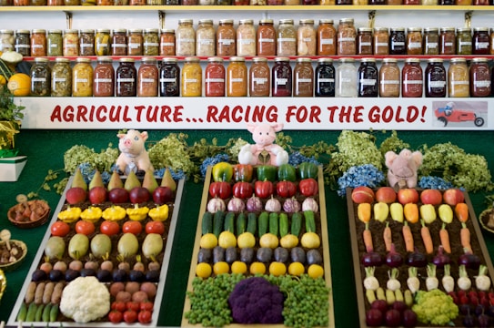 A colorful display of fruits and vegetables is organized in neat rows on three trays, surrounded by greenery and small stuffed animal figurines. Behind the display, shelves are filled with jars containing preserves and pickled goods. A banner with the text 'AGRICULTURE...RACING FOR THE GOLD!' is prominently featured.