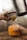 A close-up view of a hand holding a glass filled with orange juice and ice cubes. Another hand is using a metal strainer to pour fresh juice from a shaker into the glass, emphasizing a fresh beverage preparation process.
