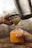 A close-up view of a hand holding a glass filled with orange juice and ice cubes. Another hand is using a metal strainer to pour fresh juice from a shaker into the glass, emphasizing a fresh beverage preparation process.
