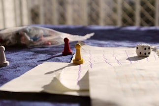 A tabletop setting includes a game board with drawn lines, scattered board game pieces such as pawns in red, yellow, and white, and a six-sided die. A blurred background with a netted pattern and diffuse lighting adds a warm, casual ambiance.
