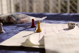 A tabletop setting includes a game board with drawn lines, scattered board game pieces such as pawns in red, yellow, and white, and a six-sided die. A blurred background with a netted pattern and diffuse lighting adds a warm, casual ambiance.