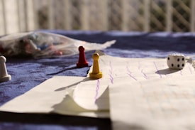 A tabletop setting includes a game board with drawn lines, scattered board game pieces such as pawns in red, yellow, and white, and a six-sided die. A blurred background with a netted pattern and diffuse lighting adds a warm, casual ambiance.